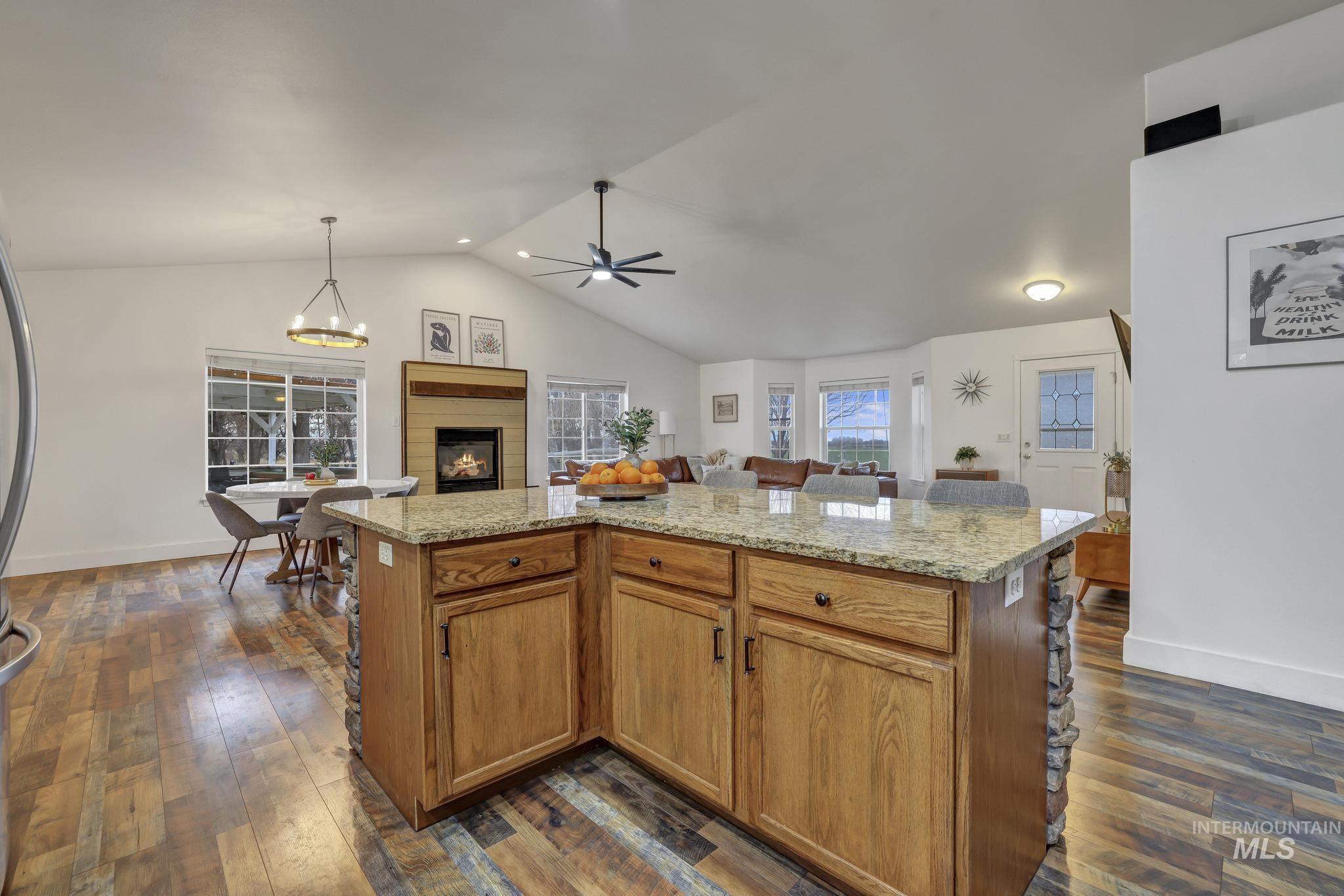 Kitchen with brown cabinets, a glass covered fireplace, light stone counters, dark wood-style flooring, and vaulted ceiling