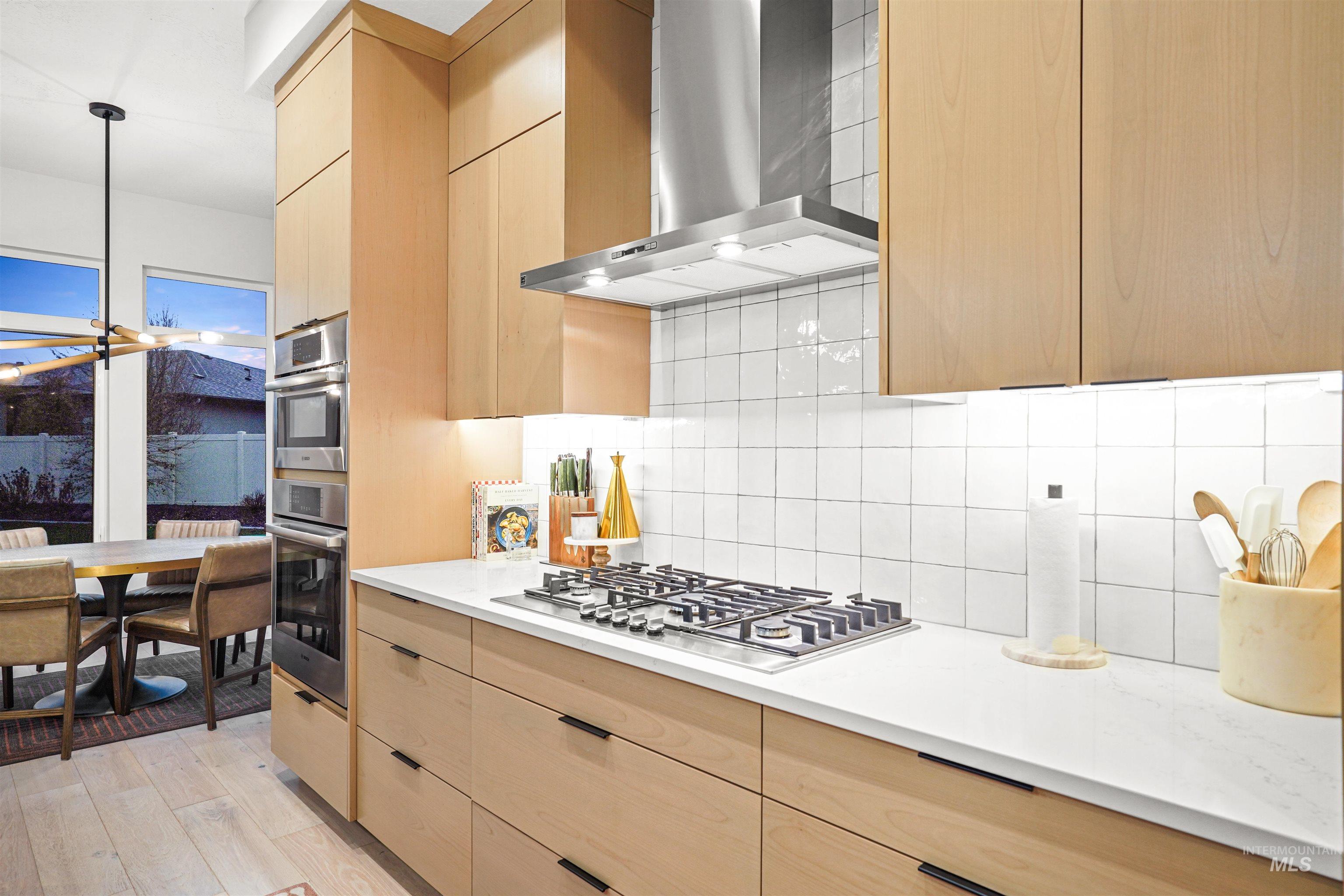 Kitchen with wall chimney range hood, light brown cabinetry, backsplash, decorative light fixtures, and light wood-style floors