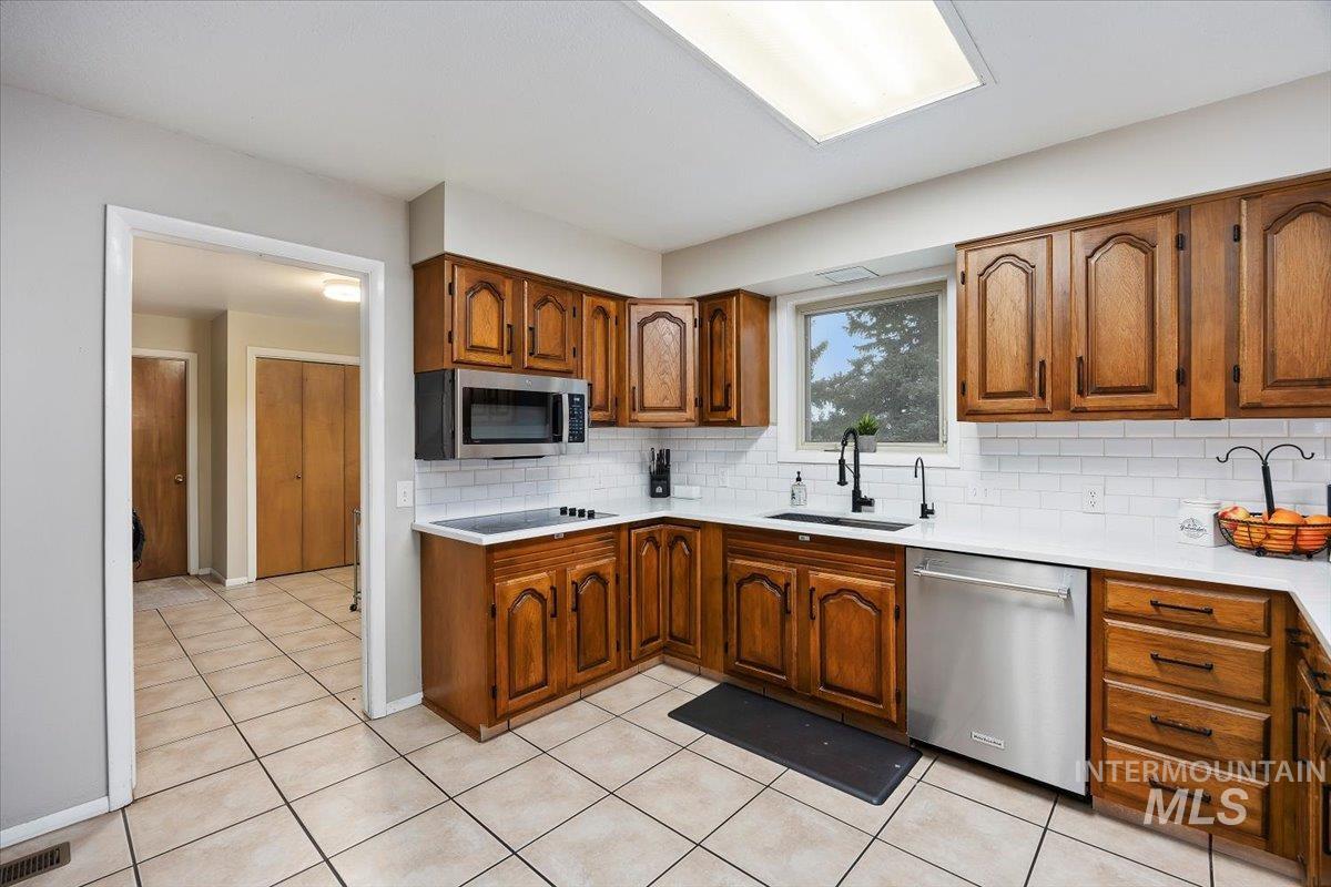 Kitchen with brown cabinetry, stainless steel appliances, decorative backsplash, light tile patterned flooring, and light stone counters