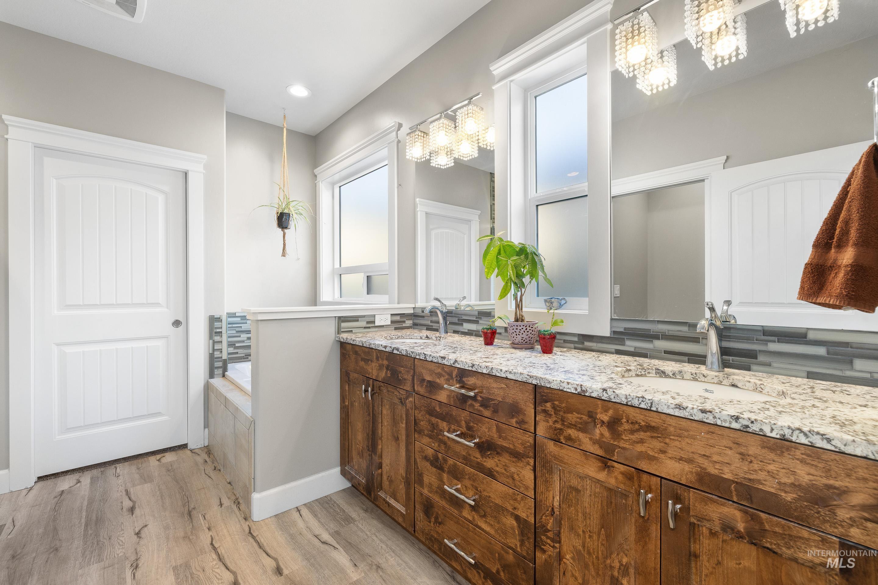 Full bath with double vanity, light wood-style floors, tasteful backsplash, and tiled tub