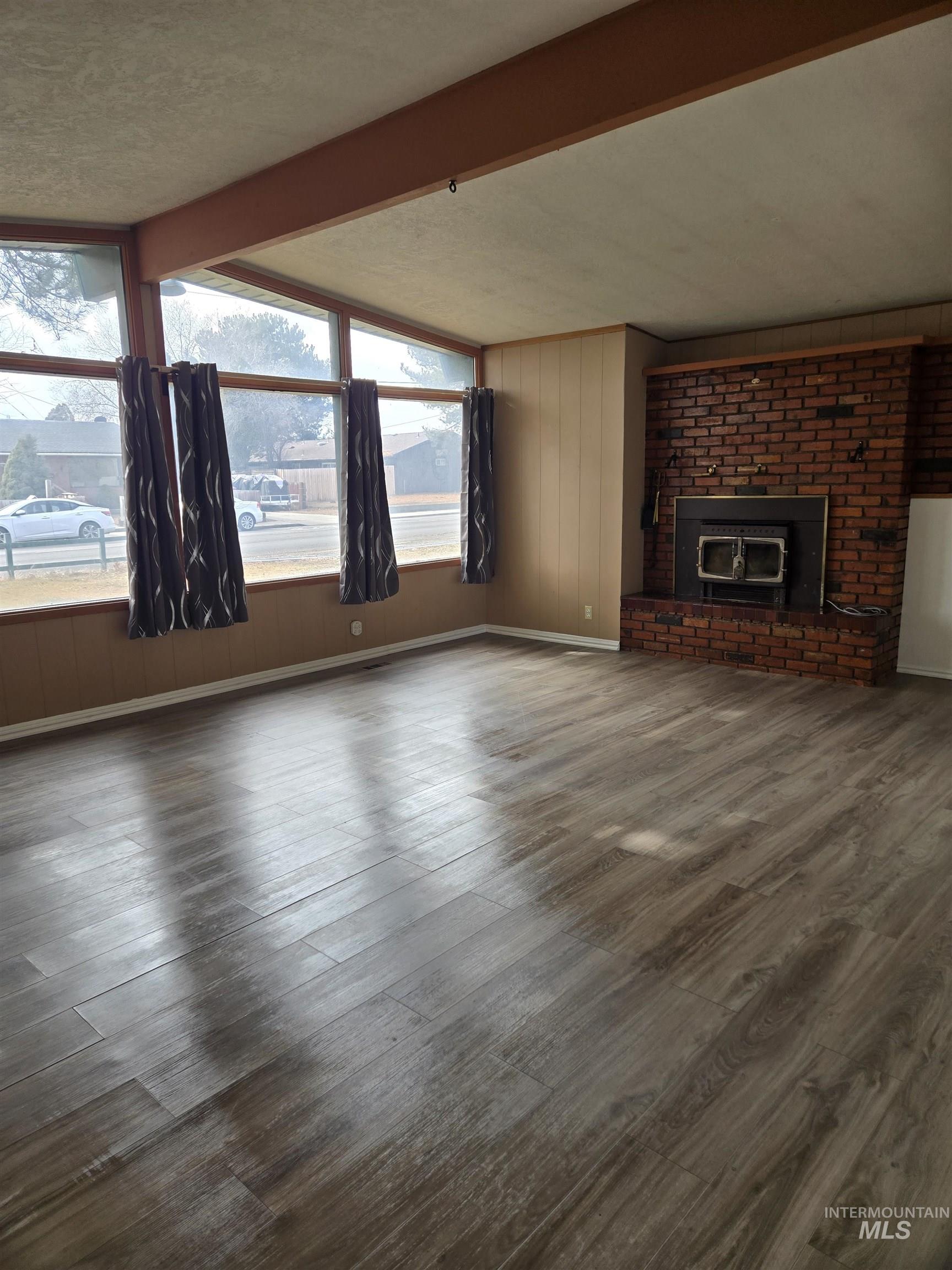 Unfurnished living room with wooden walls, beam ceiling, a brick fireplace, dark wood finished floors, and a textured ceiling