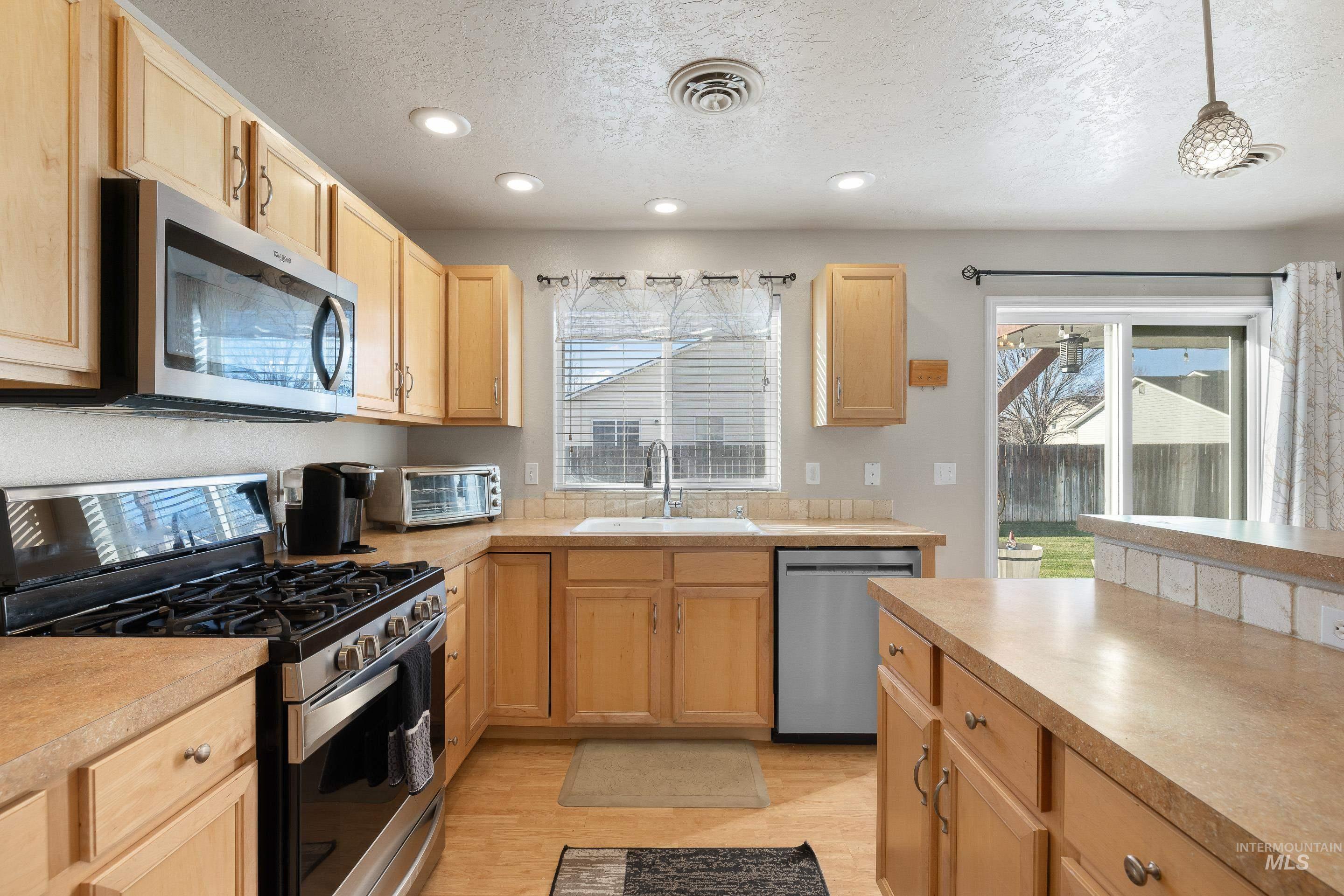 Kitchen with stainless steel appliances, light countertops, decorative light fixtures, a textured ceiling, and light brown cabinetry