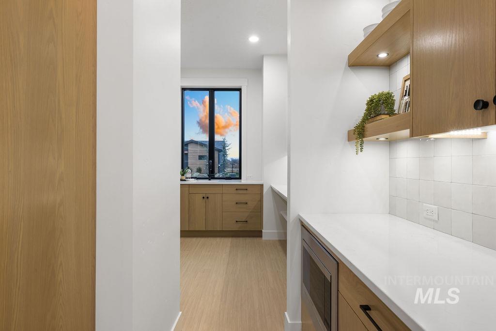 Kitchen with open shelves, brown cabinetry, light stone counters, stainless steel microwave, and light wood-style floors