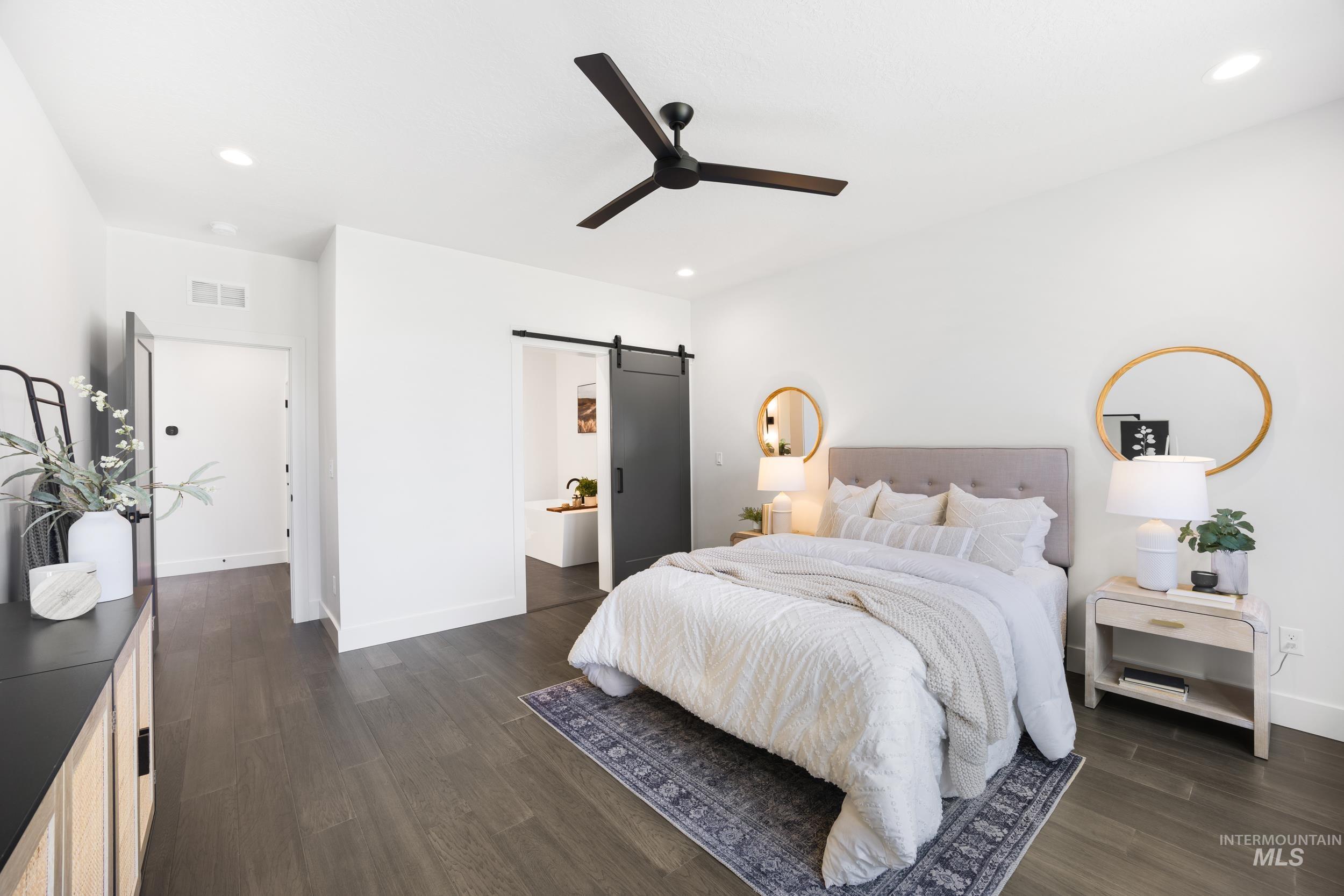 Bedroom with a barn door, dark wood finished floors, recessed lighting, ceiling fan, and ensuite bath
