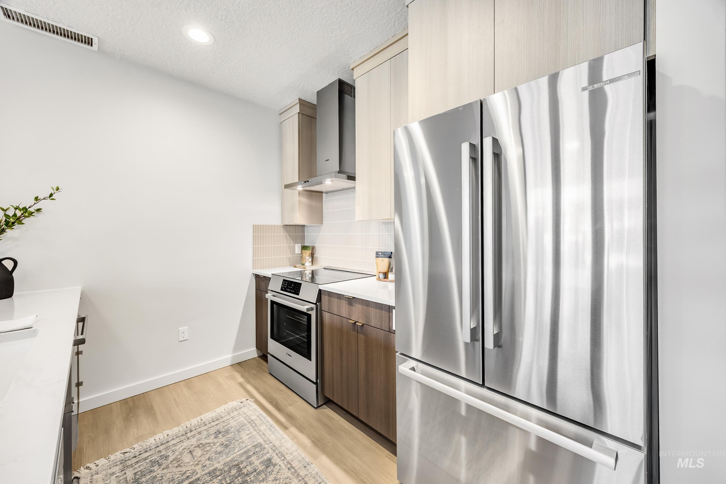 Kitchen with modern cabinets, appliances with stainless steel finishes, light wood-style flooring, wall chimney exhaust hood, and a textured ceiling