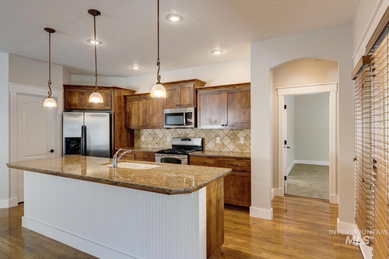 Kitchen with brown cabinets, light stone counters, appliances with stainless steel finishes, decorative backsplash, and pendant lighting
