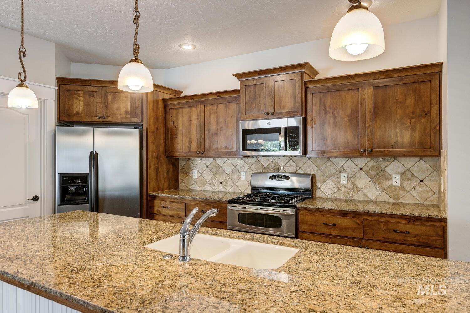 Kitchen with brown cabinetry, appliances with stainless steel finishes, decorative light fixtures, light stone countertops, and a textured ceiling