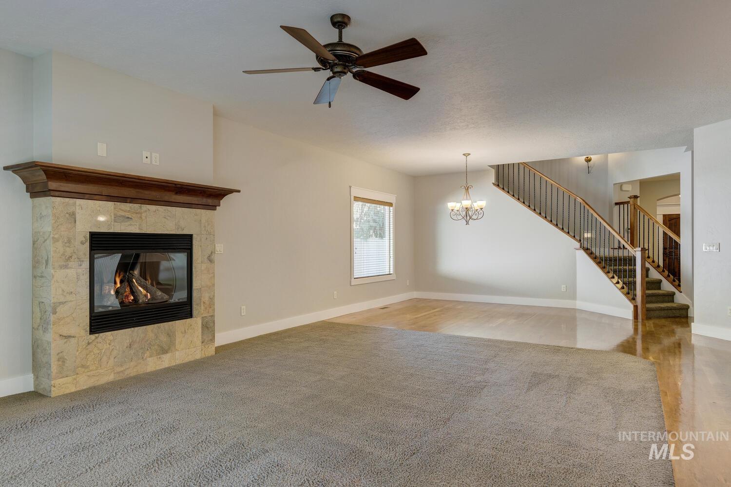 Unfurnished living room featuring a tiled fireplace, stairway, a ceiling fan, a chandelier, and light colored carpet