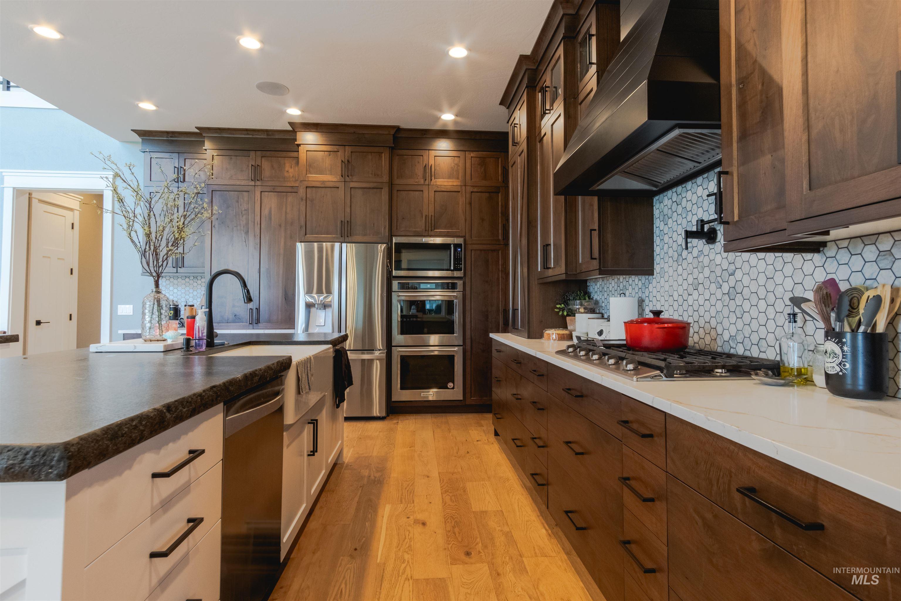 Kitchen featuring custom range hood, stainless steel appliances, backsplash, light wood-style floors, and dark brown cabinetry