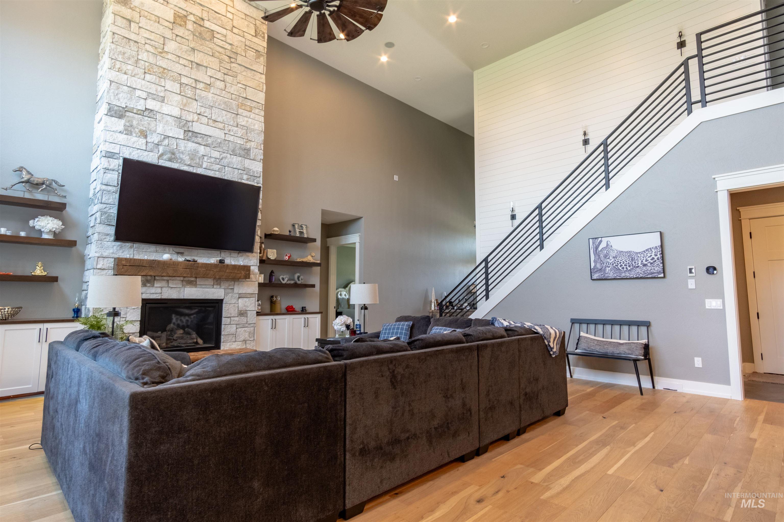 Living room featuring a high ceiling, light wood-style floors, a stone fireplace, ceiling fan, and stairs