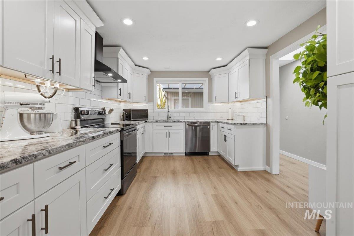Kitchen with stainless steel appliances, white cabinets, light stone countertops, wall chimney exhaust hood, and light wood-style floors