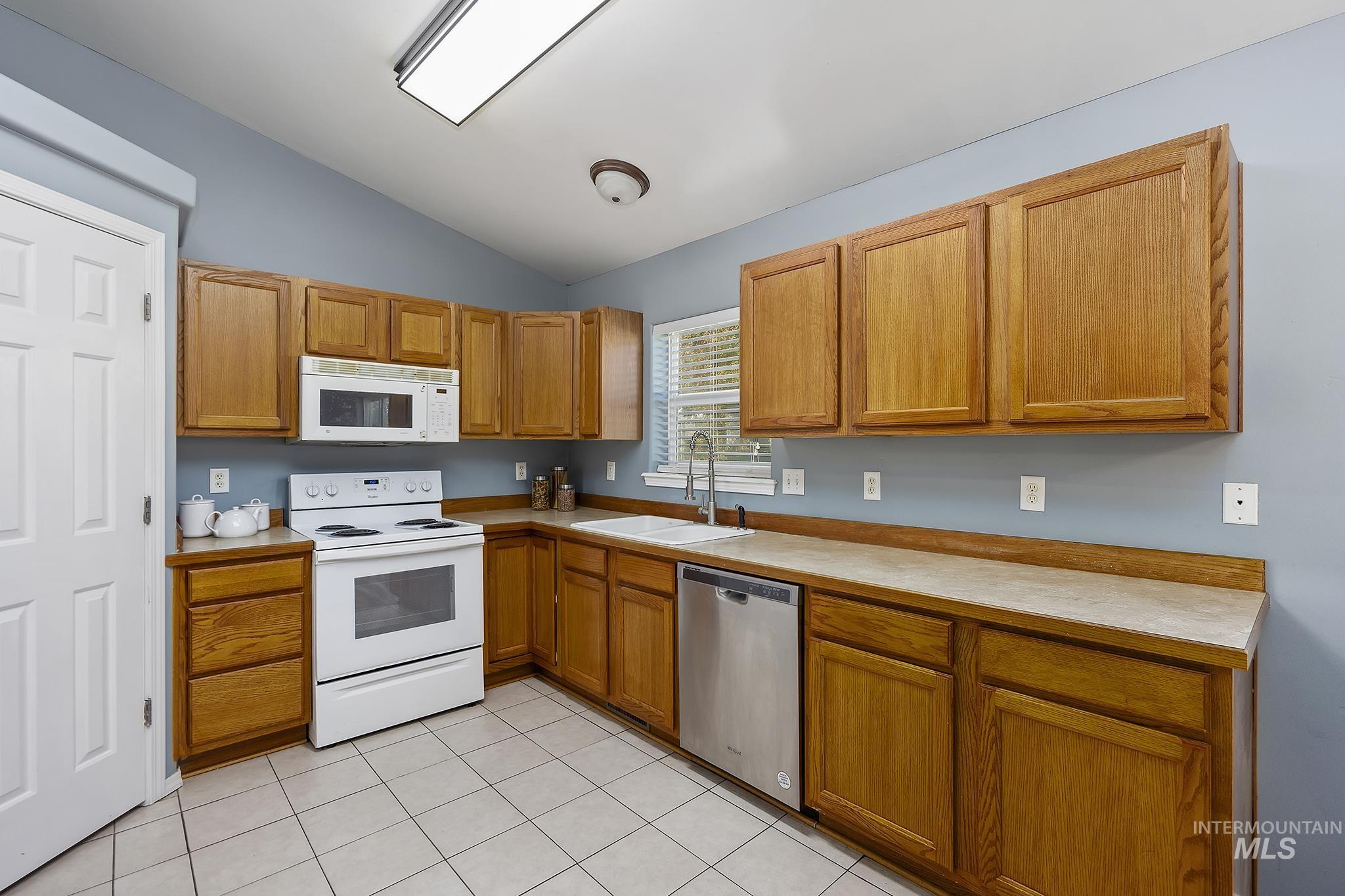 Kitchen featuring white appliances, light countertops, lofted ceiling, brown cabinetry, and light tile patterned floors