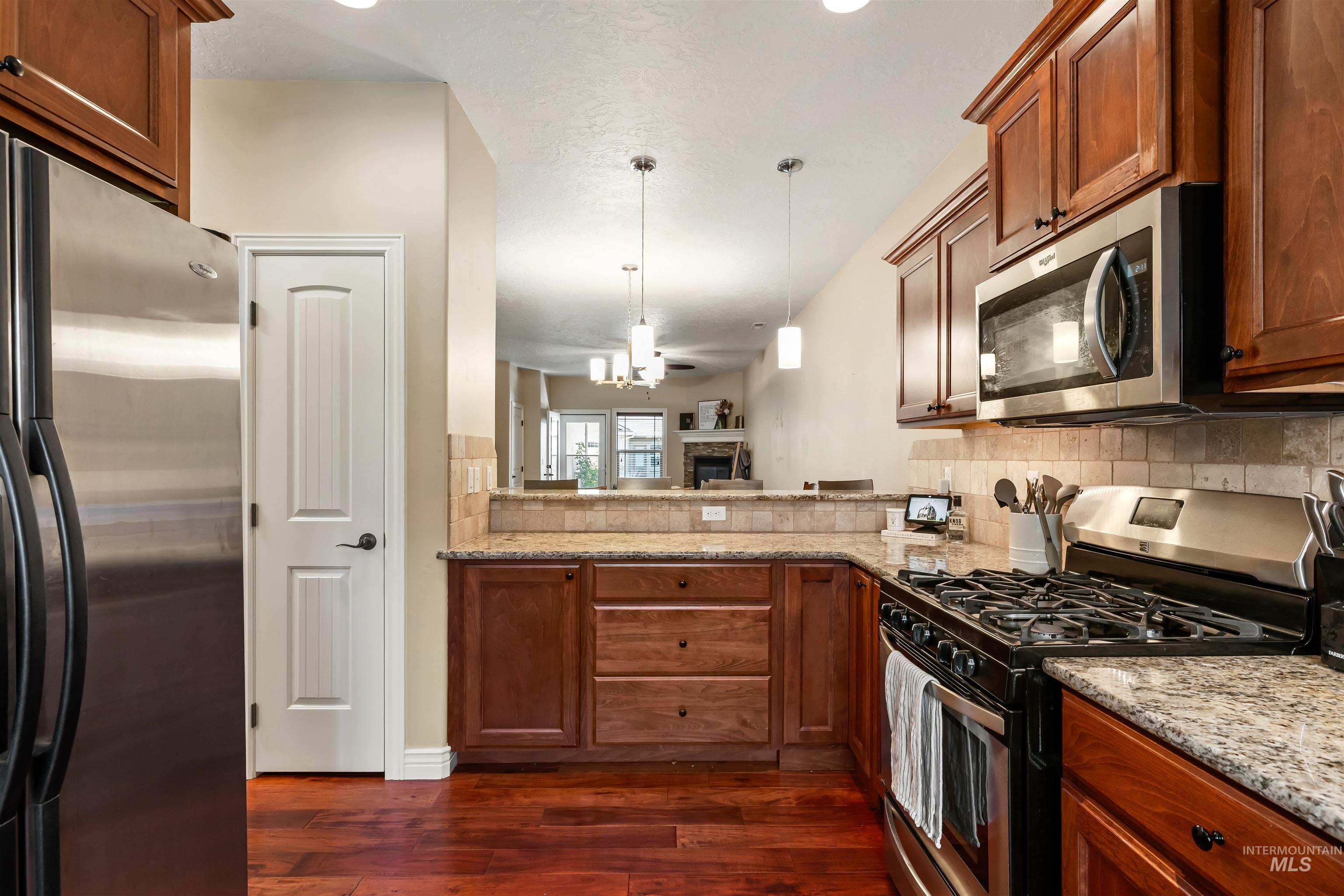 Kitchen with stainless steel appliances, hanging light fixtures, brown cabinetry, and light stone countertops
