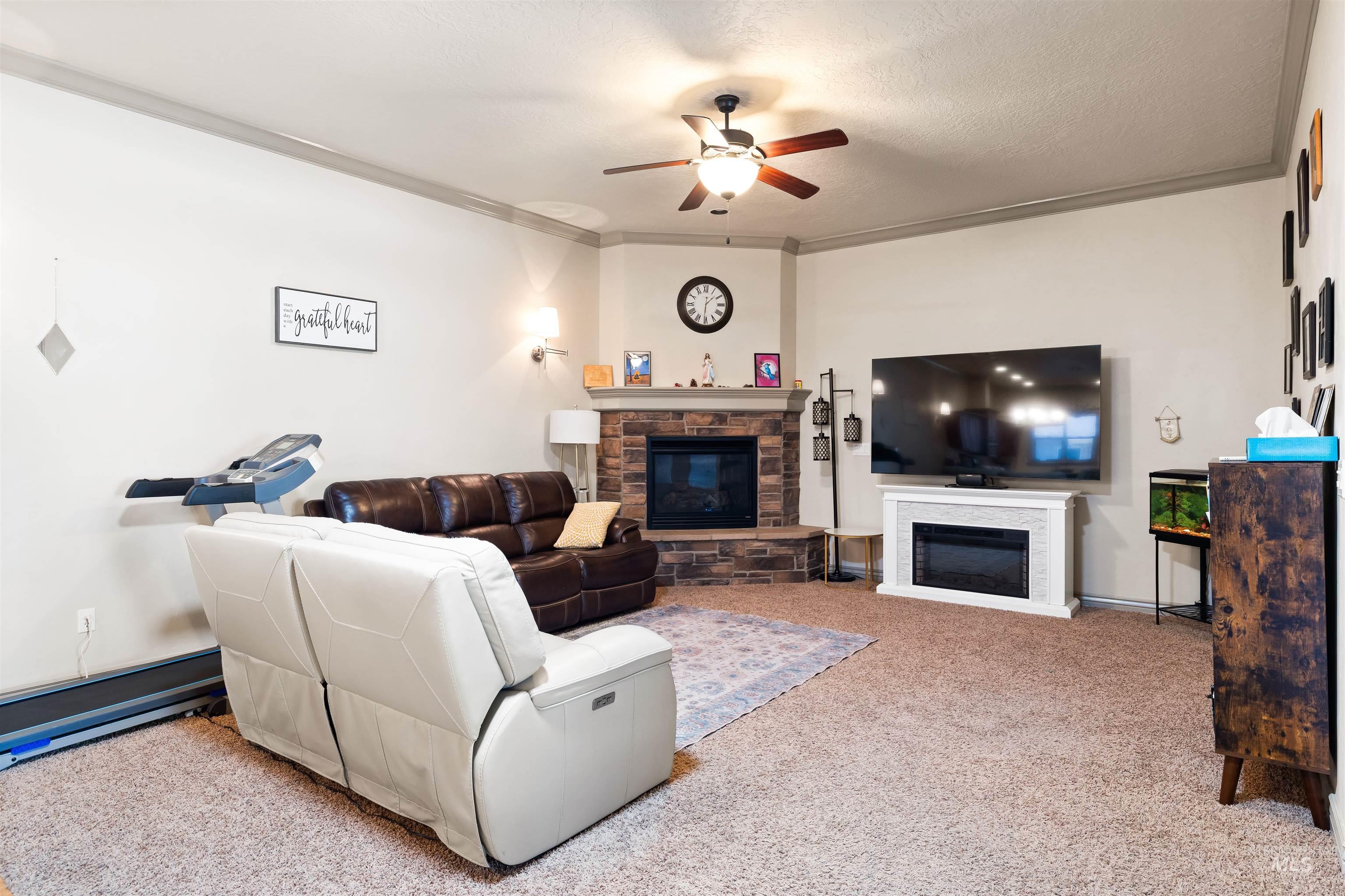 Living room featuring a stone fireplace, carpet, a ceiling fan, ornamental molding, and a textured ceiling