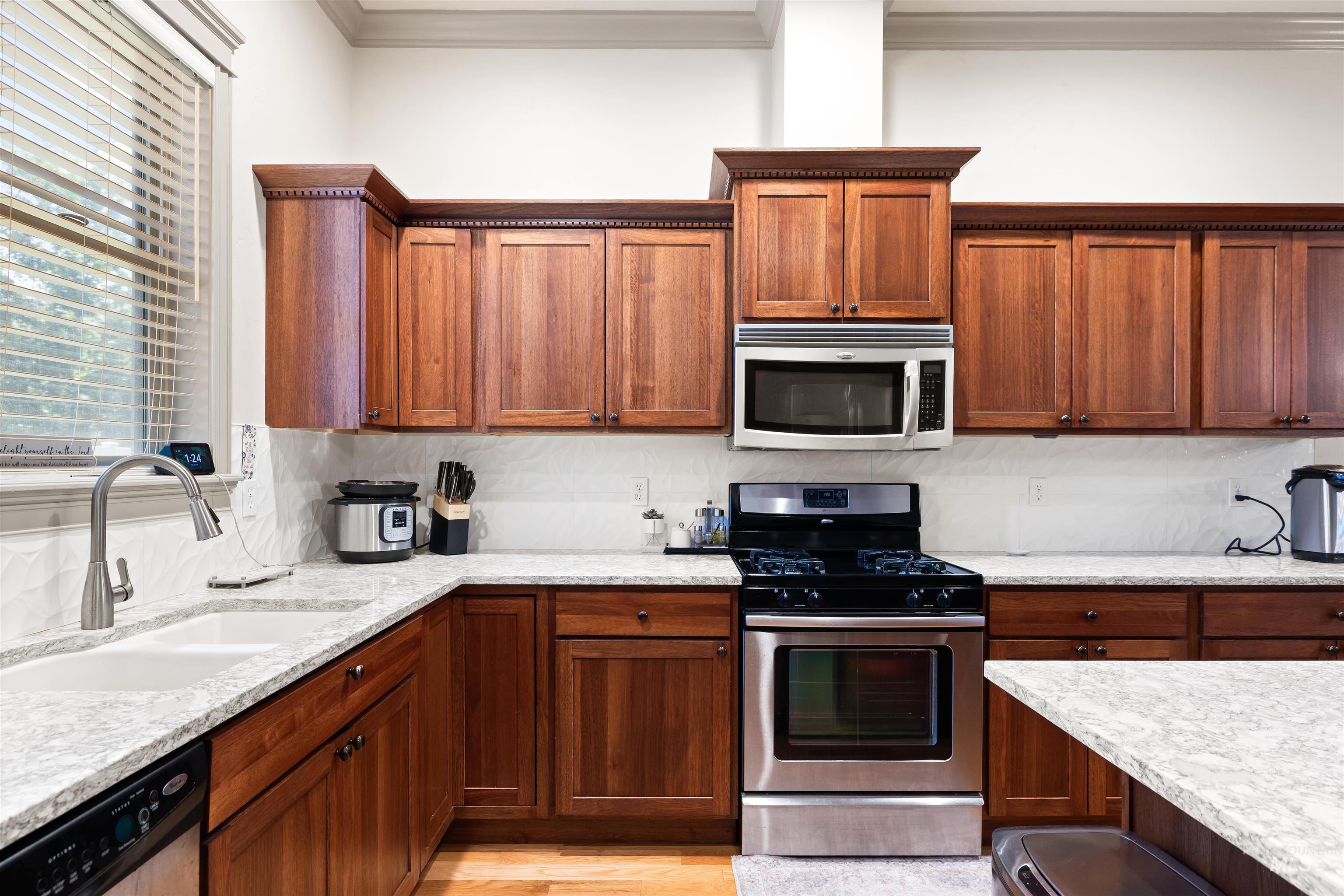 Kitchen featuring stainless steel appliances, brown cabinetry, light stone counters, ornamental molding, and decorative backsplash