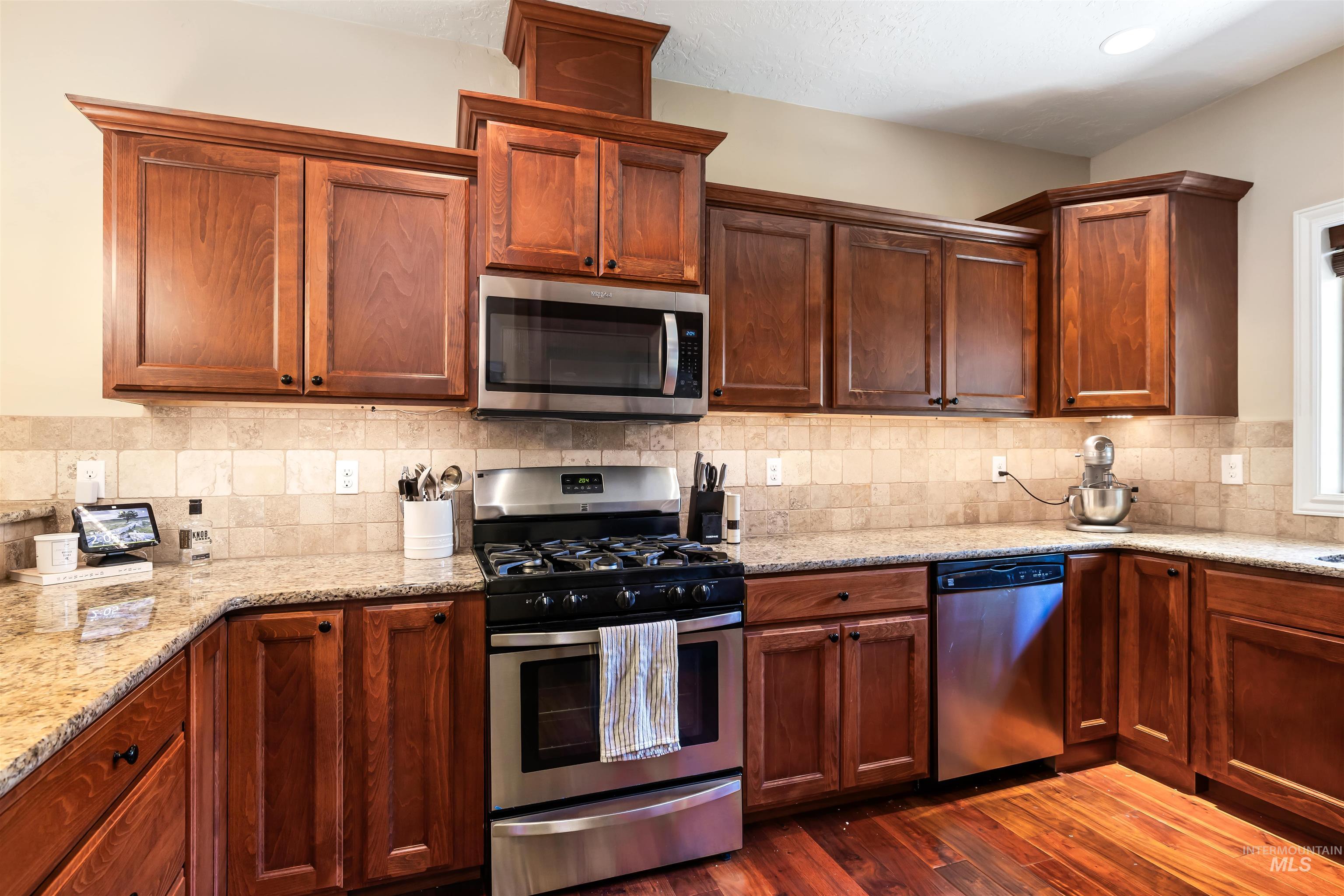 Kitchen featuring stainless steel appliances, light stone counters, dark wood-style floors, tasteful backsplash, and brown cabinetry