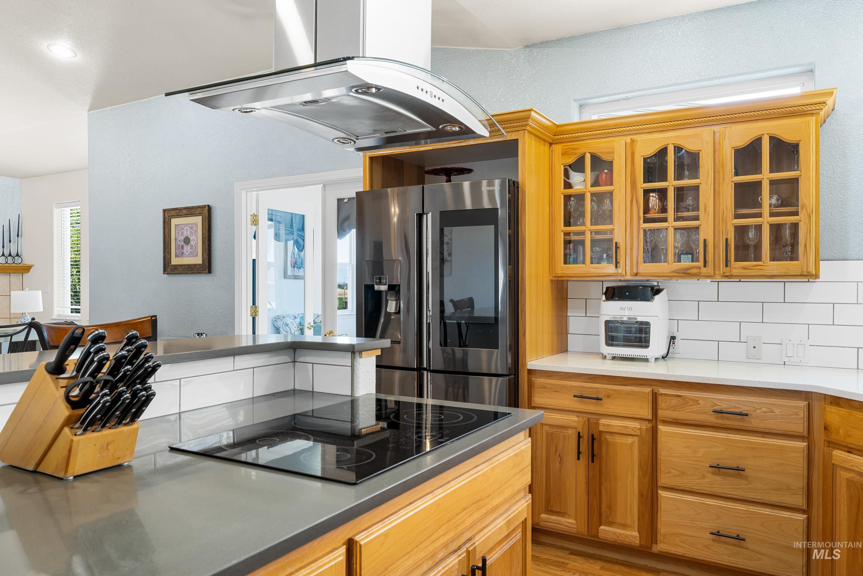 Kitchen with glass insert cabinets, decorative backsplash, stainless steel fridge with ice dispenser, island range hood, and black electric cooktop
