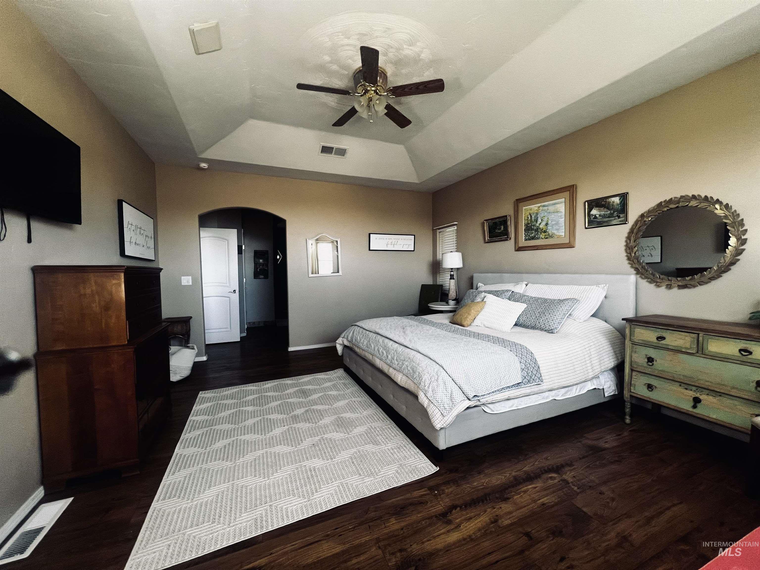 Bedroom with a raised ceiling, arched walkways, dark wood-style floors, and a ceiling fan