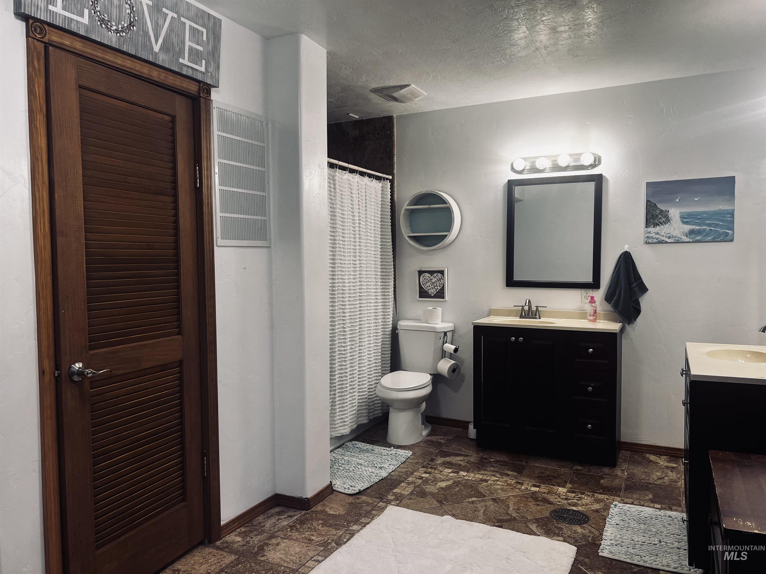 Full bath featuring a shower with curtain, two vanities, a textured ceiling, and stone tile flooring