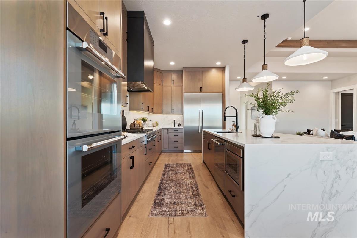 Kitchen with built in appliances, light stone countertops, decorative light fixtures, light wood-style flooring, and a center island with sink