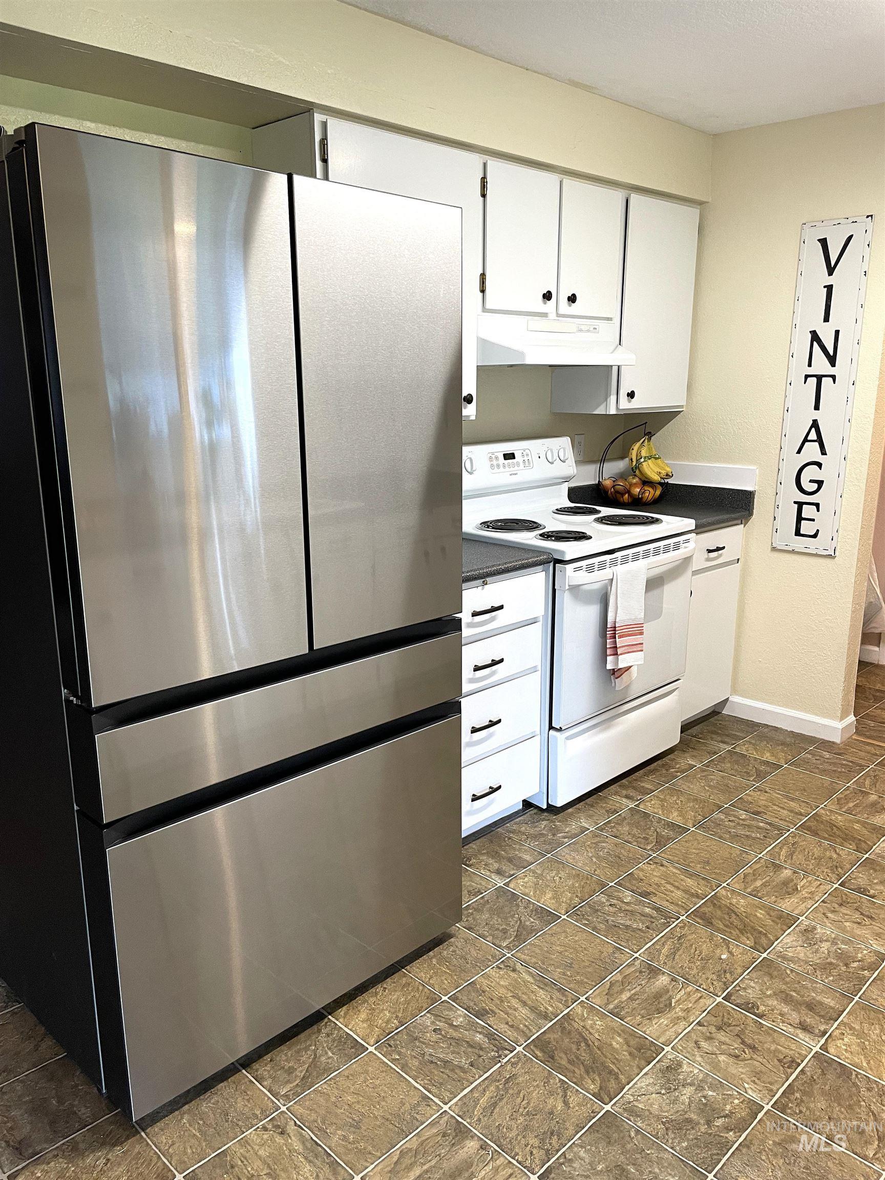 Kitchen with freestanding refrigerator, electric range, white cabinetry, dark countertops, and under cabinet range hood