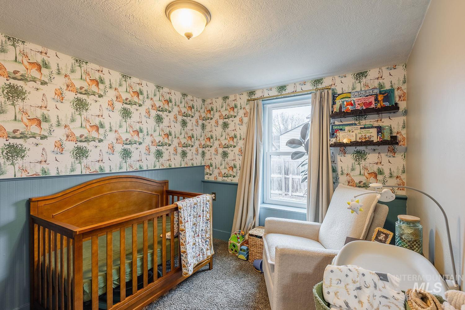 Bedroom featuring wallpapered walls, a textured ceiling, wainscoting, a crib, and carpet
