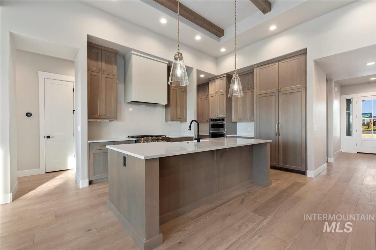 Kitchen featuring a kitchen island with sink, premium range hood, decorative light fixtures, light wood-style floors, and light stone countertops