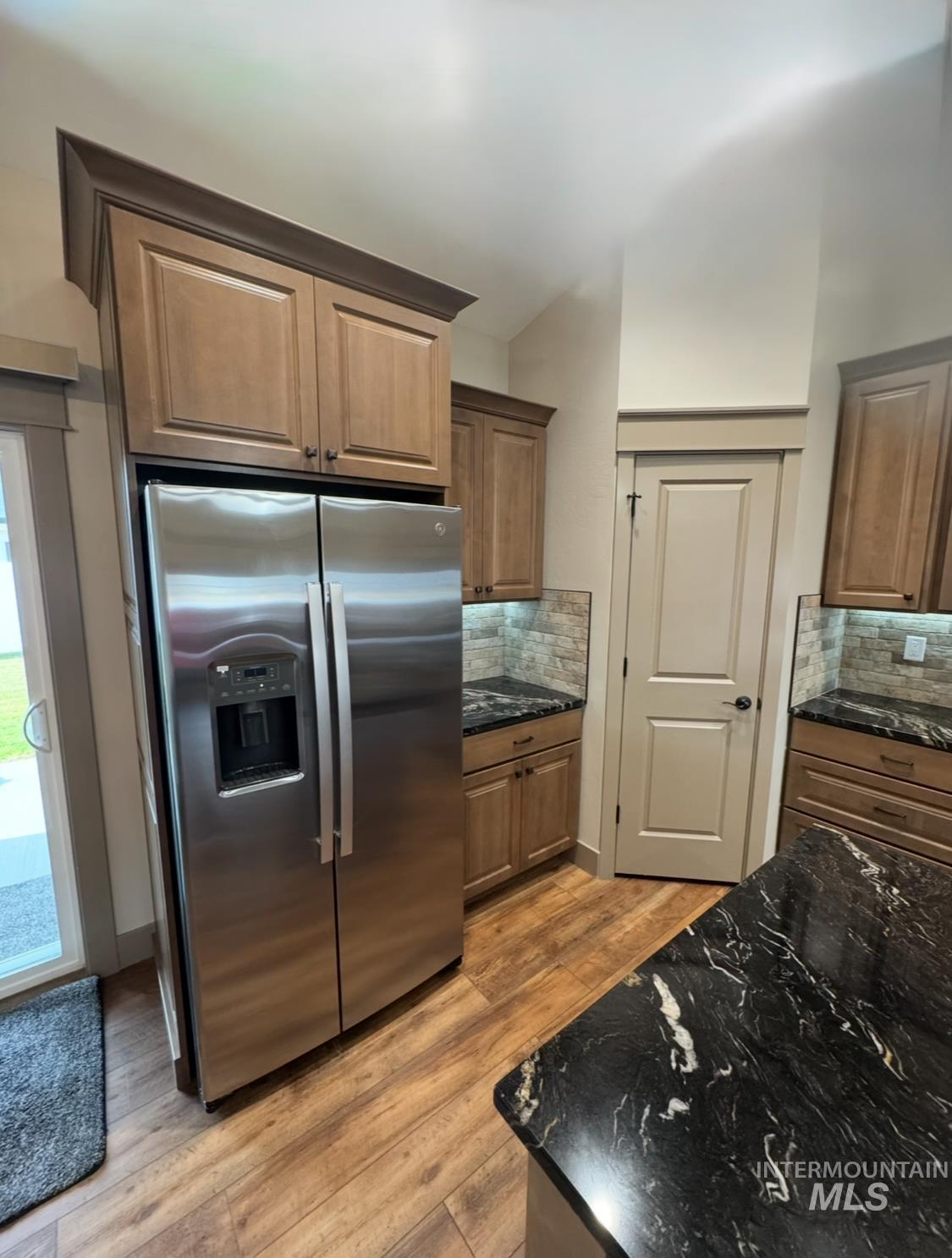 Kitchen with stainless steel fridge, dark stone countertops, decorative backsplash, light wood finished floors, and brown cabinetry