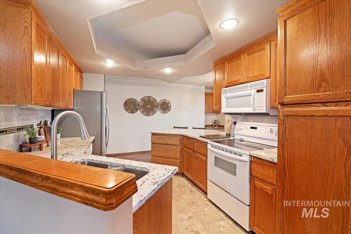 Kitchen with a peninsula, white appliances, a tray ceiling, light stone counters, and brown cabinetry