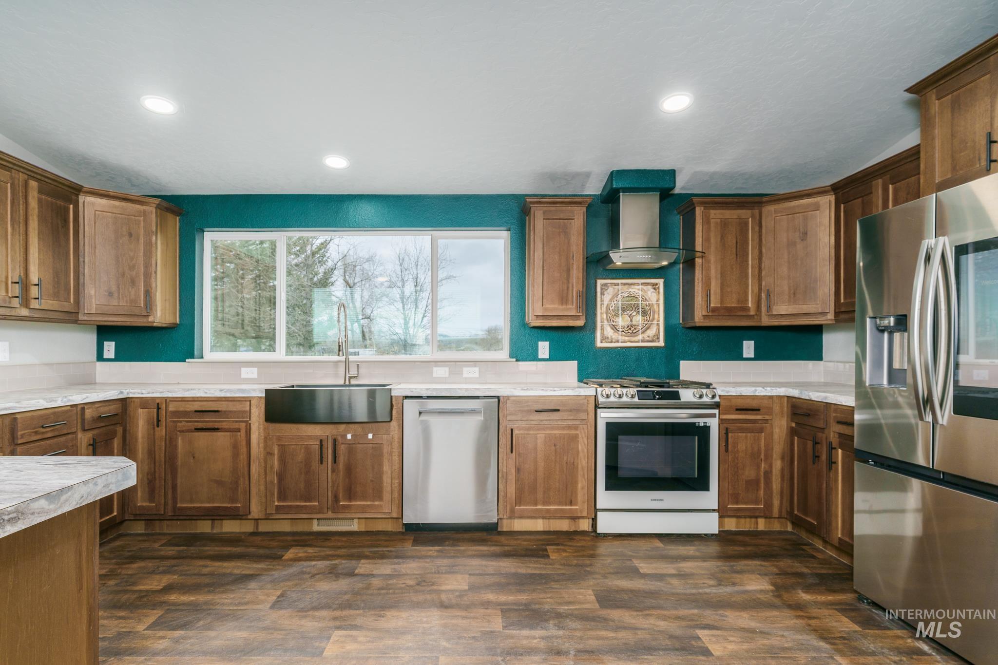 Kitchen featuring appliances with stainless steel finishes, wall chimney range hood, light countertops, brown cabinetry, and dark wood-type flooring