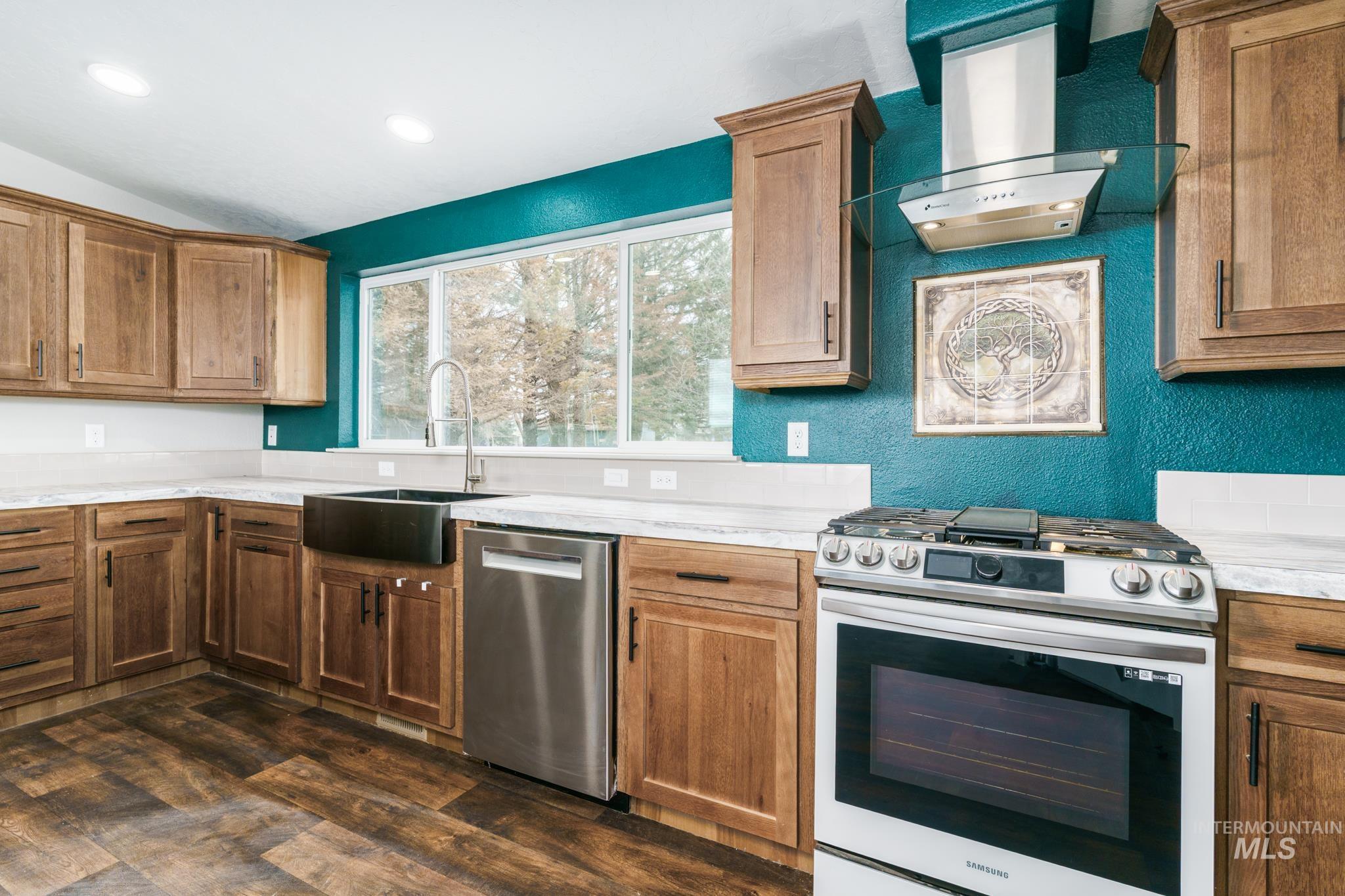 Kitchen featuring appliances with stainless steel finishes, wall chimney exhaust hood, a textured wall, brown cabinets, and vaulted ceiling