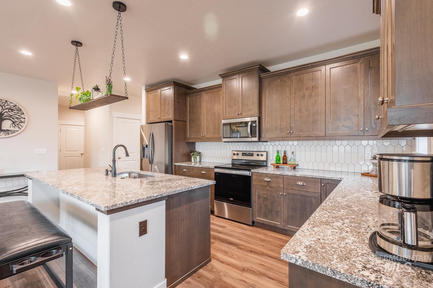 Kitchen with appliances with stainless steel finishes, an island with sink, light stone countertops, decorative light fixtures, and light wood-type flooring