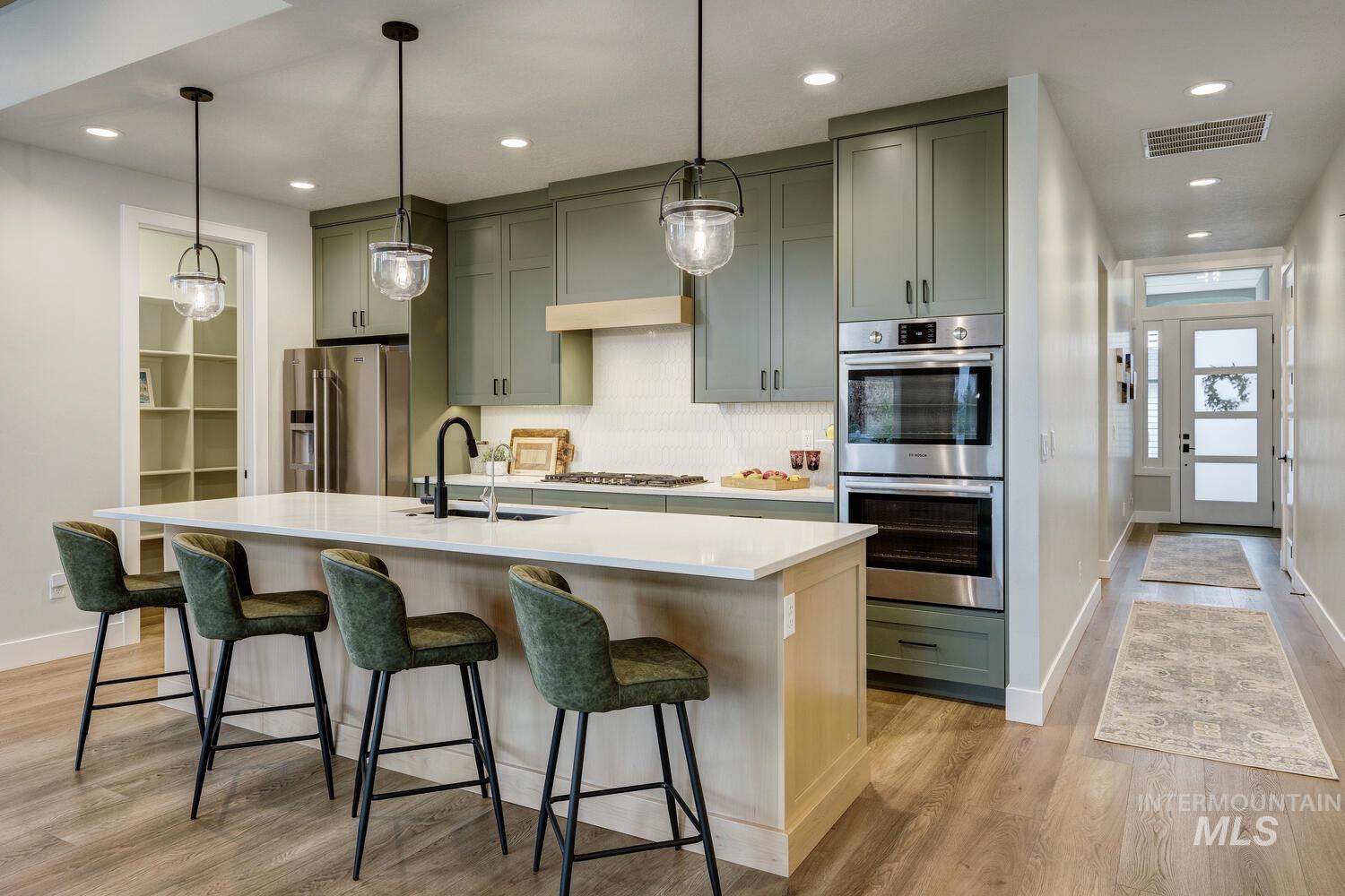 Kitchen featuring green cabinets, appliances with stainless steel finishes, decorative light fixtures, a center island with sink, and light stone counters