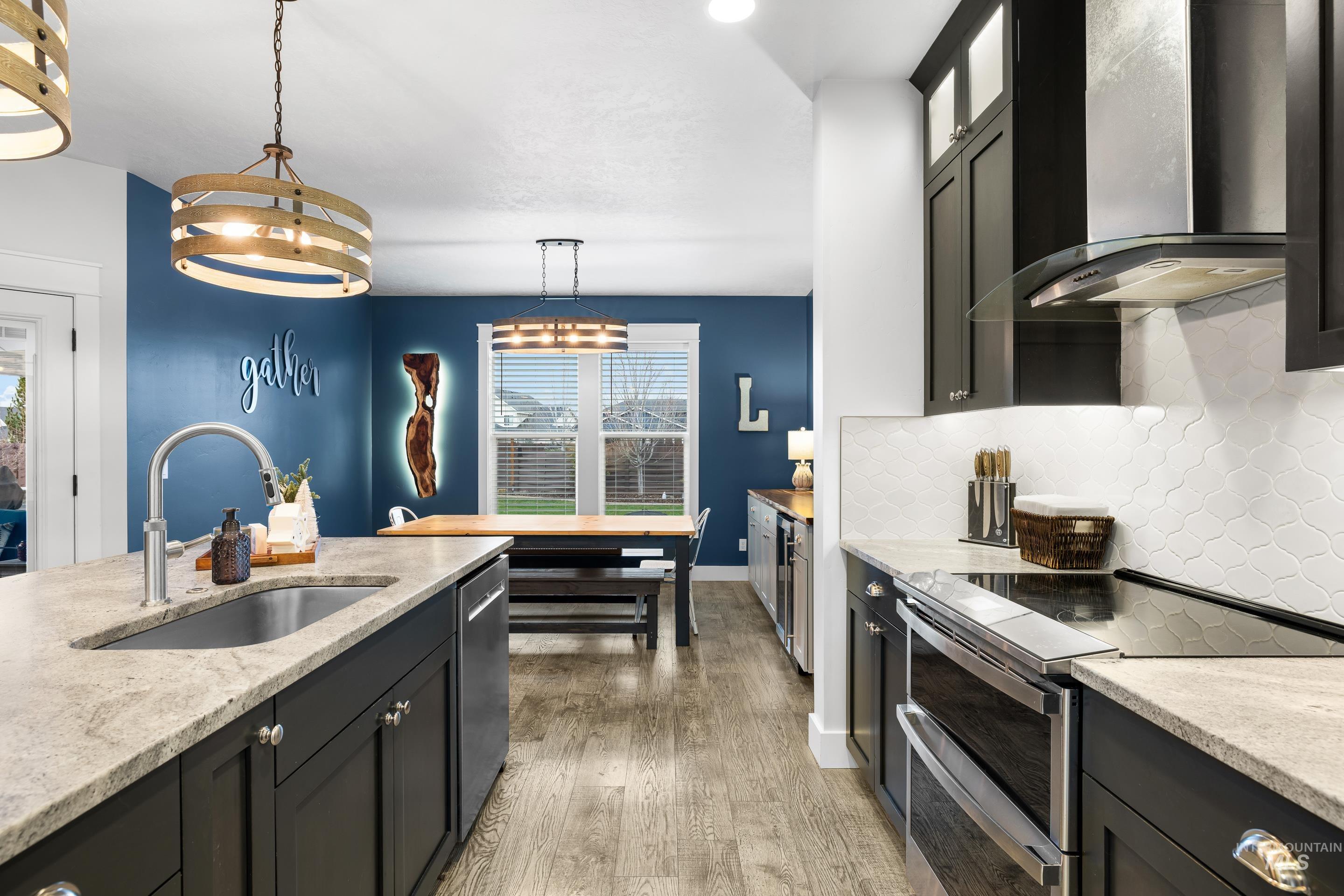 Kitchen with dark cabinetry, stainless steel appliances, wall chimney range hood, light stone countertops, and hanging light fixtures