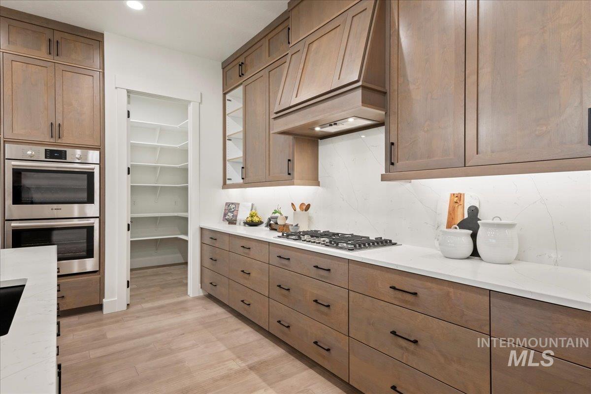 Kitchen with appliances with stainless steel finishes, custom exhaust hood, light wood-style floors, tasteful backsplash, and brown cabinetry