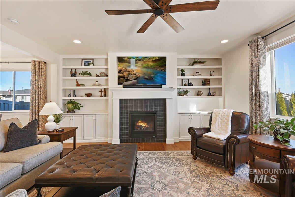 Living room with a tiled fireplace, ceiling fan, wood finished floors, and built in features