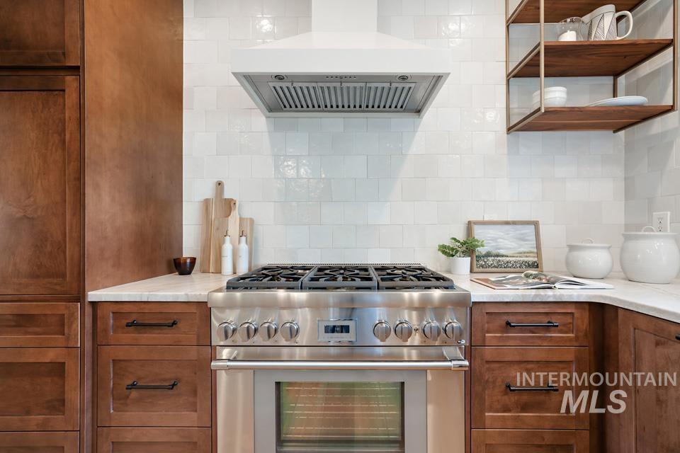 Kitchen featuring high end stainless steel range oven, ventilation hood, tasteful backsplash, open shelves, and light stone countertops