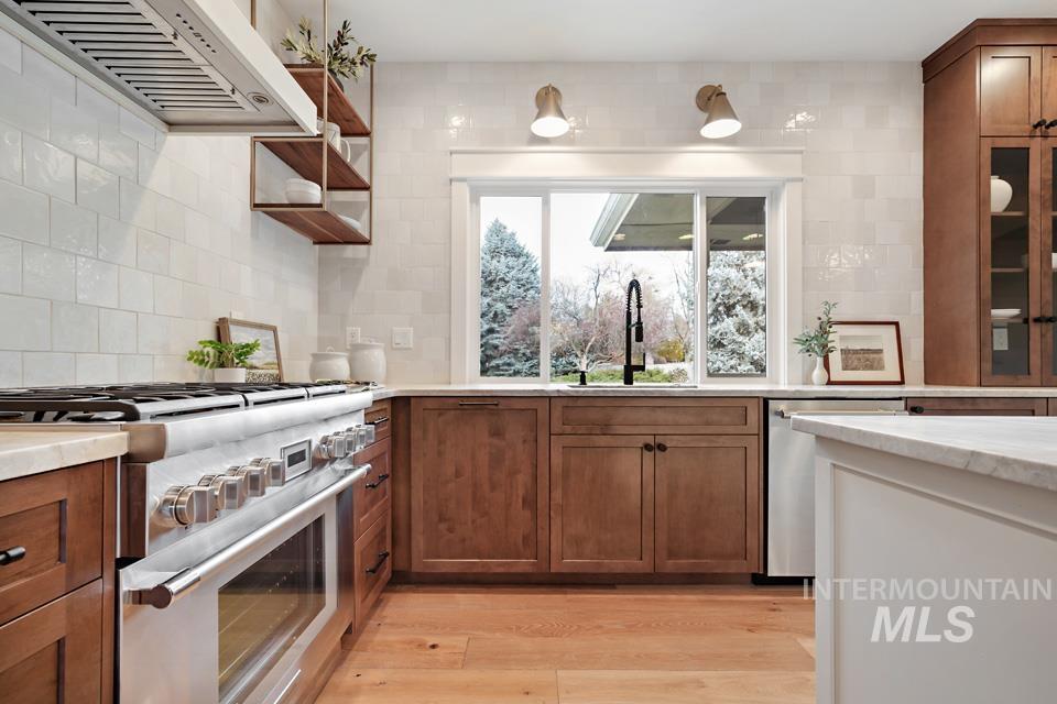Kitchen featuring stainless steel appliances, custom range hood, brown cabinets, light wood finished floors, and open shelves