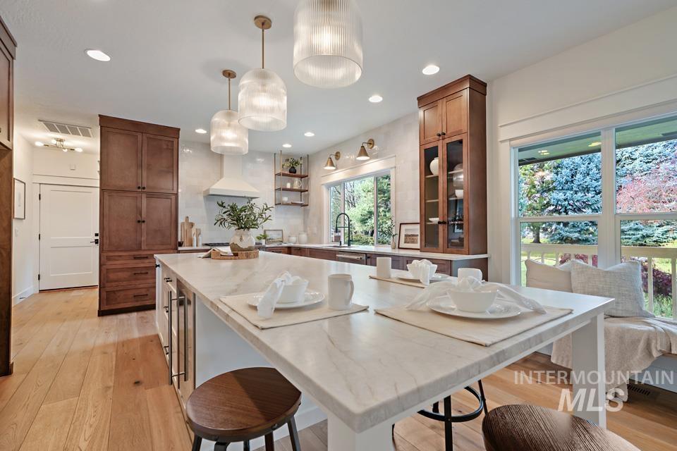 Kitchen with hanging light fixtures, light wood-style flooring, brown cabinetry, light stone counters, and open shelves