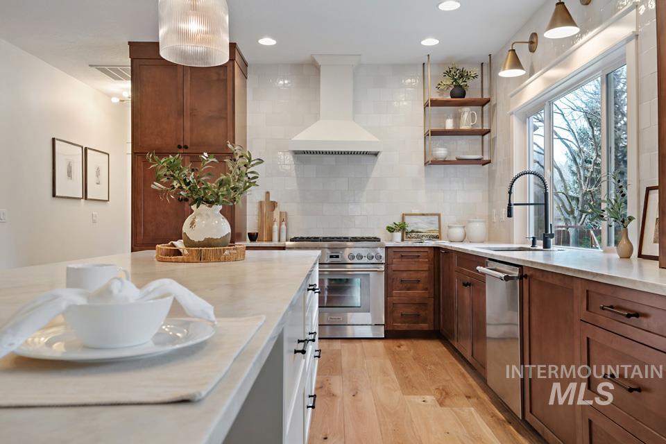 Kitchen featuring custom exhaust hood, appliances with stainless steel finishes, light wood-style flooring, brown cabinets, and open shelves