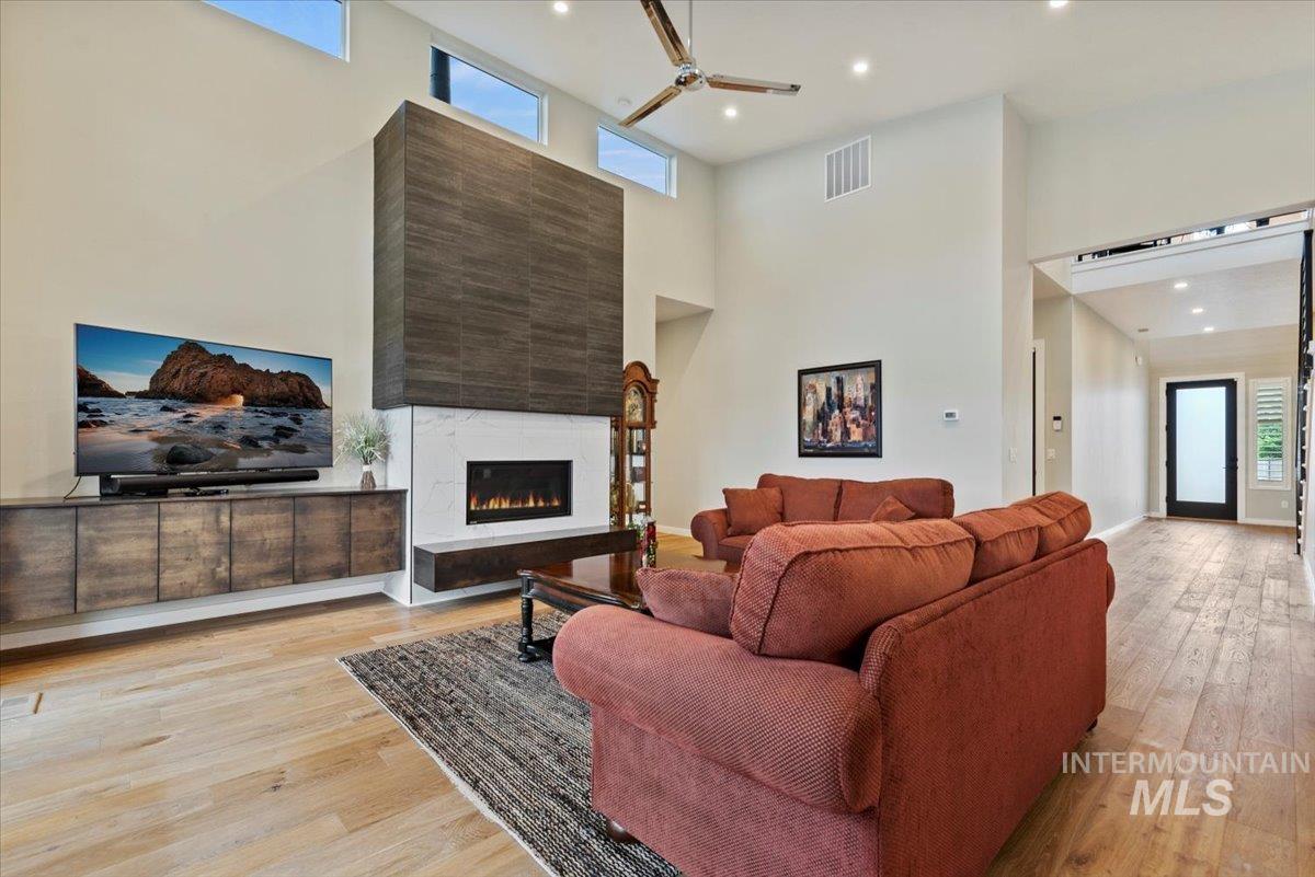 Living room with light wood-style flooring, a glass covered fireplace, recessed lighting, a towering ceiling, and a ceiling fan