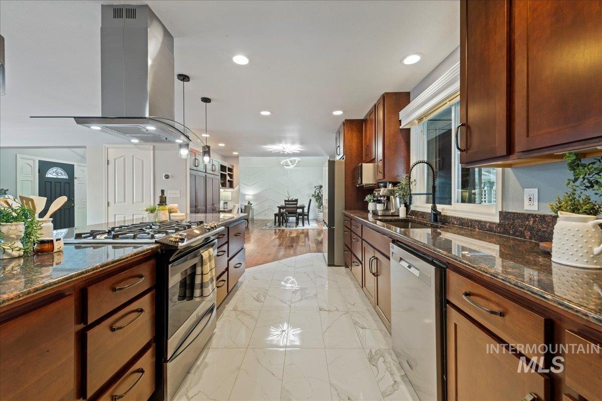 Kitchen featuring dark stone counters, appliances with stainless steel finishes, island range hood, pendant lighting, and light marble finish floors