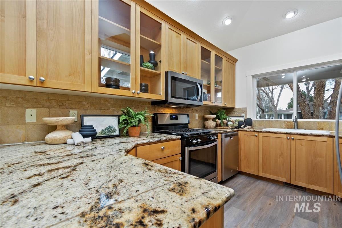 Kitchen featuring appliances with stainless steel finishes, glass insert cabinets, backsplash, dark wood-type flooring, and light stone countertops