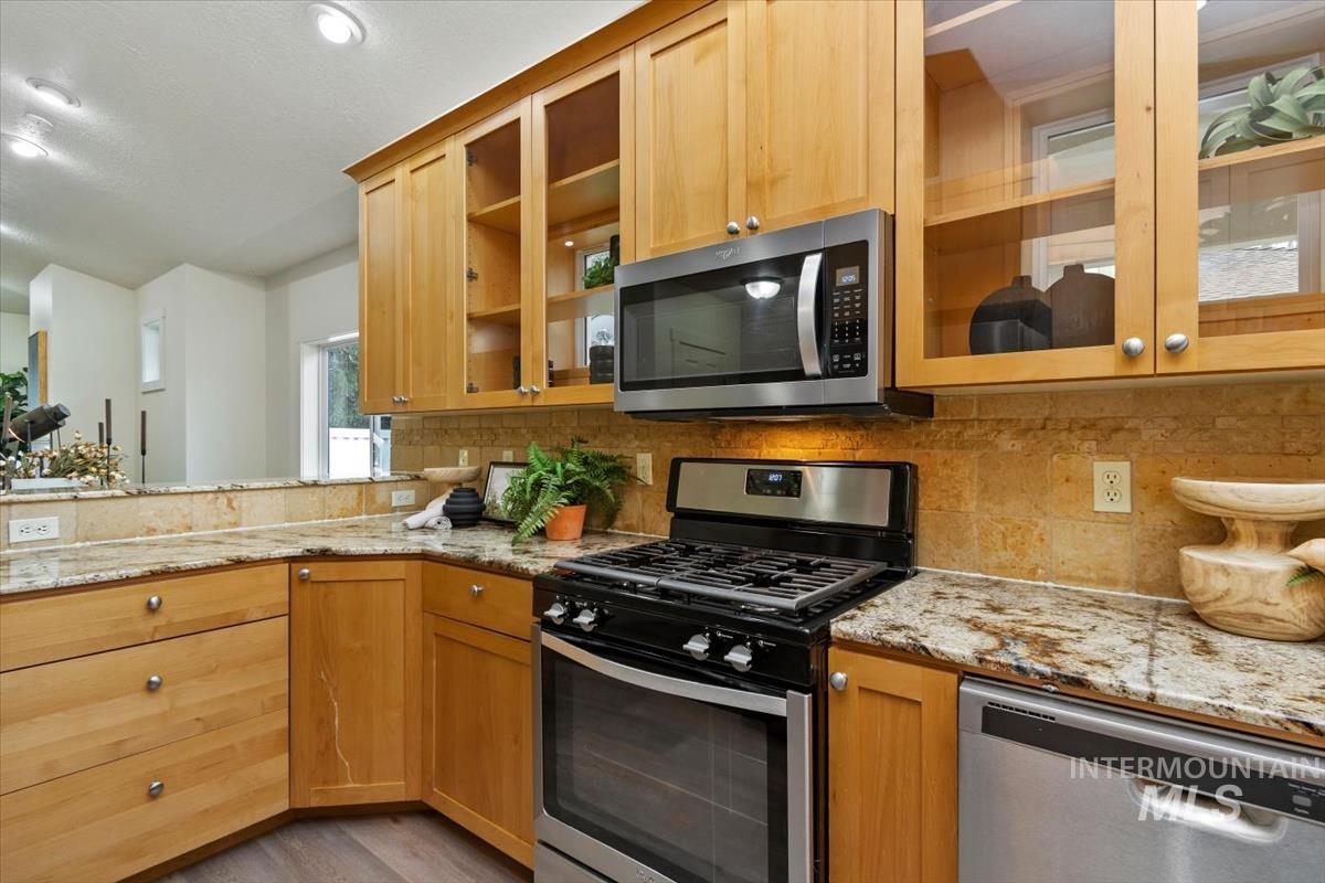 Kitchen featuring appliances with stainless steel finishes, glass insert cabinets, light stone counters, decorative backsplash, and recessed lighting