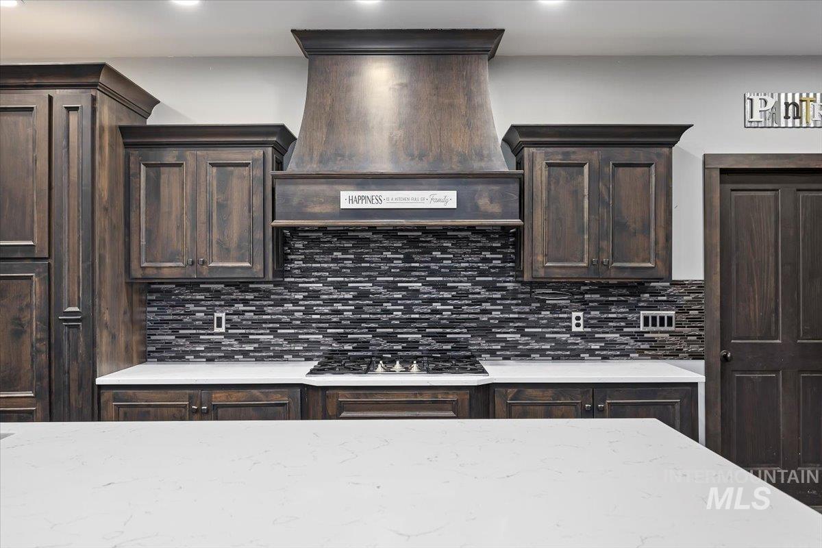 Kitchen featuring dark brown cabinetry, custom exhaust hood, light stone countertops, and backsplash