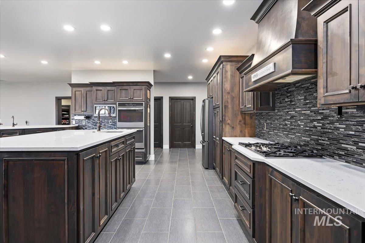 Kitchen featuring dark brown cabinetry, light stone countertops, custom range hood, backsplash, and recessed lighting