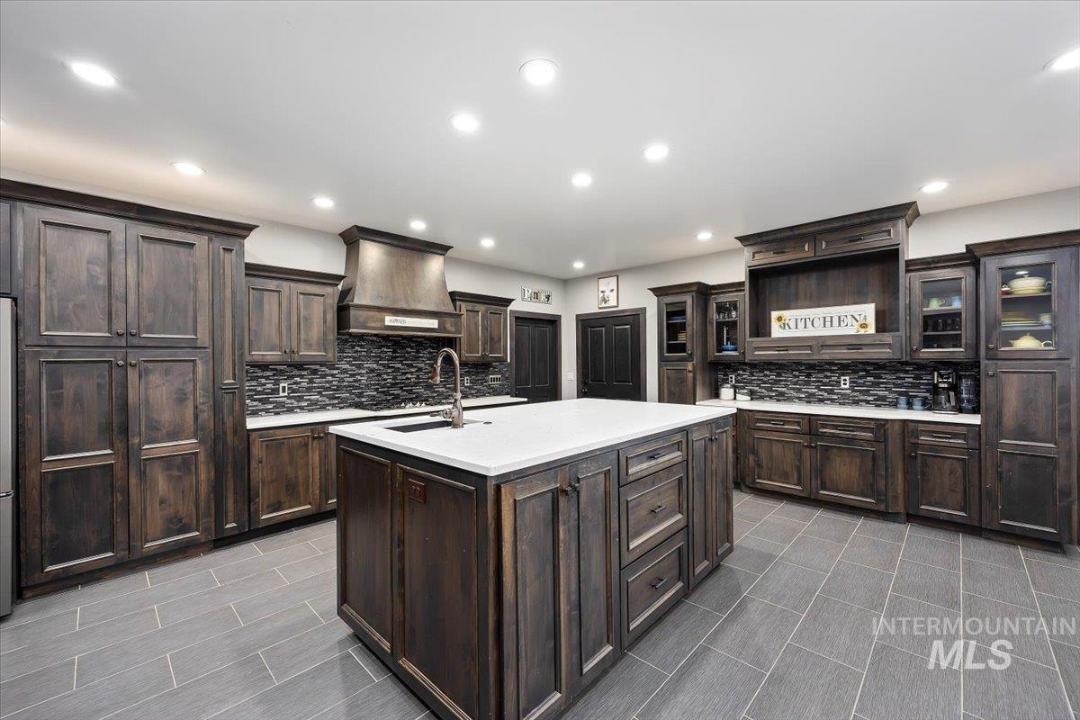 Kitchen featuring dark brown cabinets, a center island with sink, glass insert cabinets, custom exhaust hood, and light stone countertops