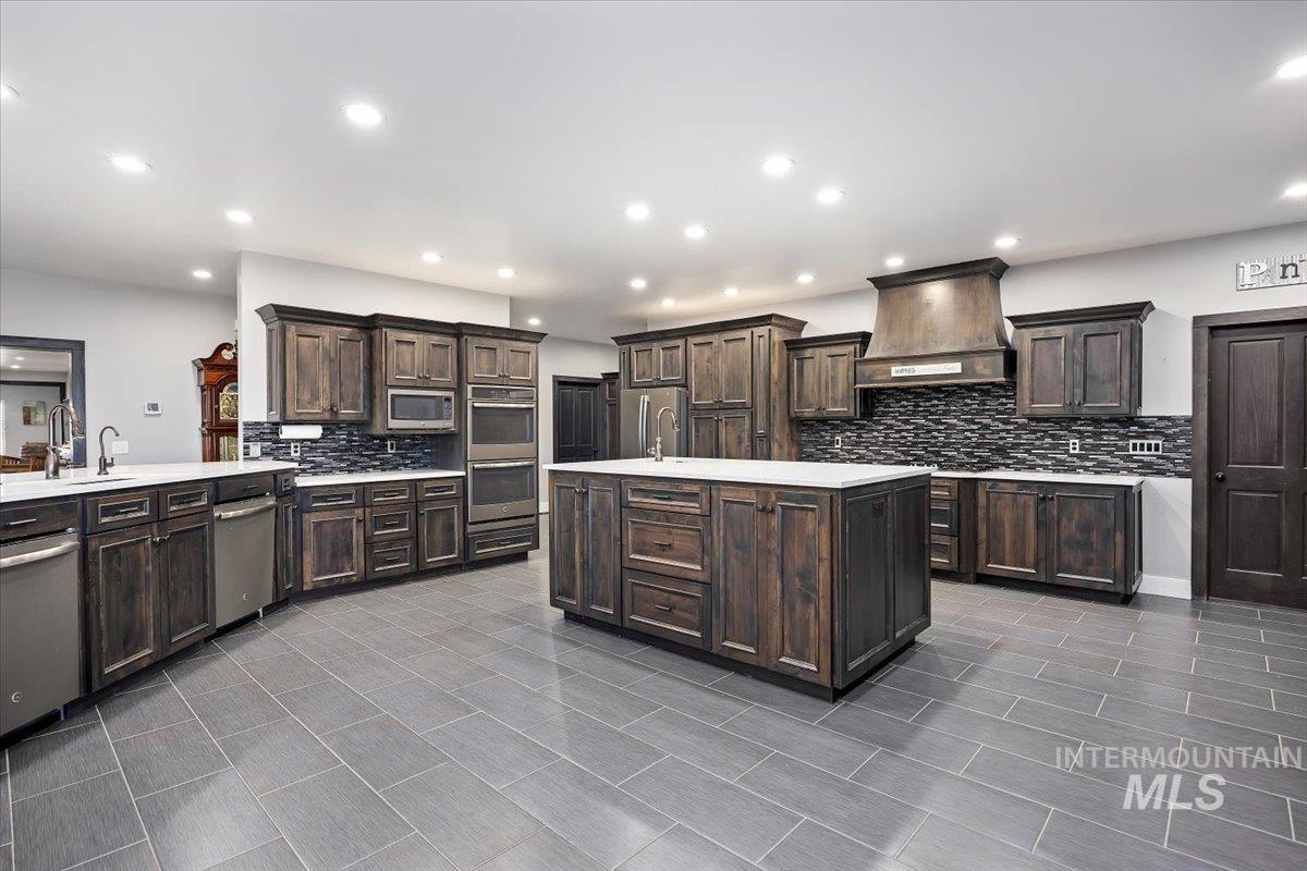 Kitchen with dark brown cabinets, custom range hood, backsplash, a kitchen island with sink, and recessed lighting