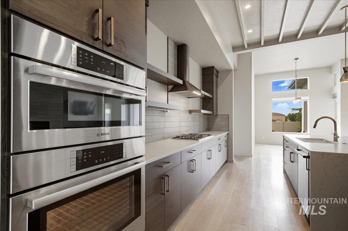 Kitchen featuring stainless steel appliances, light wood-style flooring, wall chimney range hood, backsplash, and pendant lighting