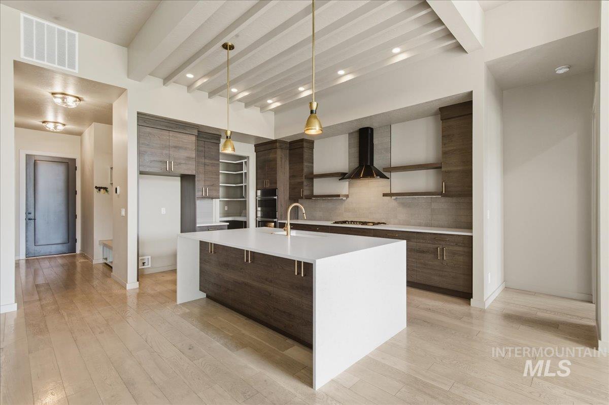 Kitchen featuring modern cabinets, open shelves, beamed ceiling, a center island with sink, and light wood finished floors